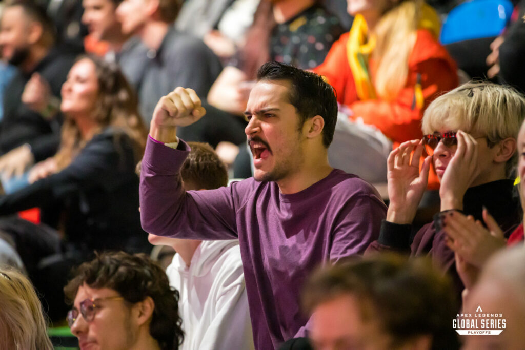 A fan wearing a purple T shirt cheers for his favorite team from the stands. Surrounding him are dozens of fans seated with various levels of excitement.