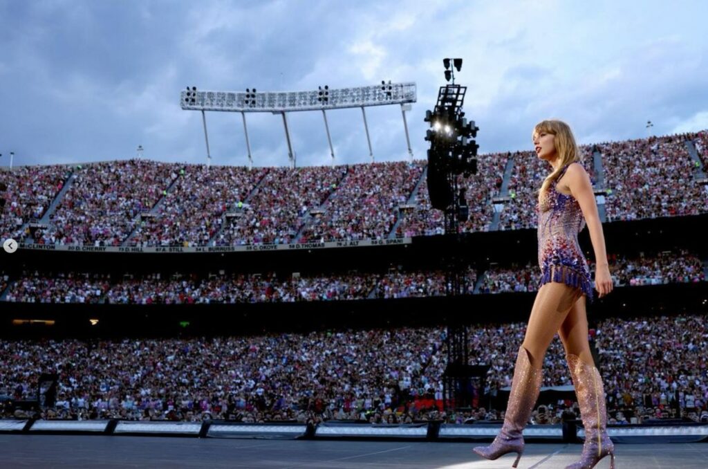 Taylor Swift walking on to the stage. In the background is a packed stadium with thousands of fans.