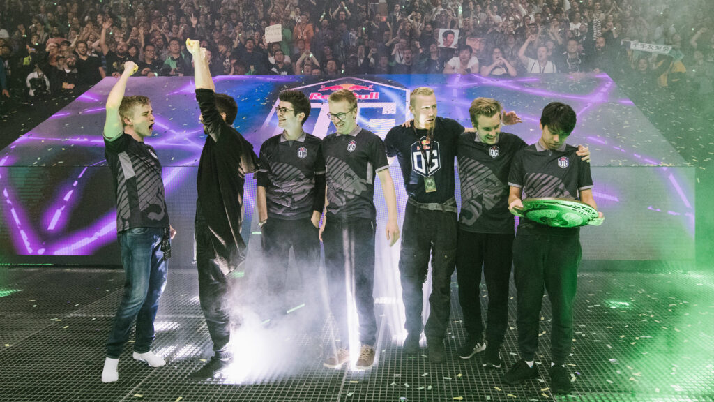 OG with their hands raised on-stage at TI 2018, while ana looks at the Aegis.