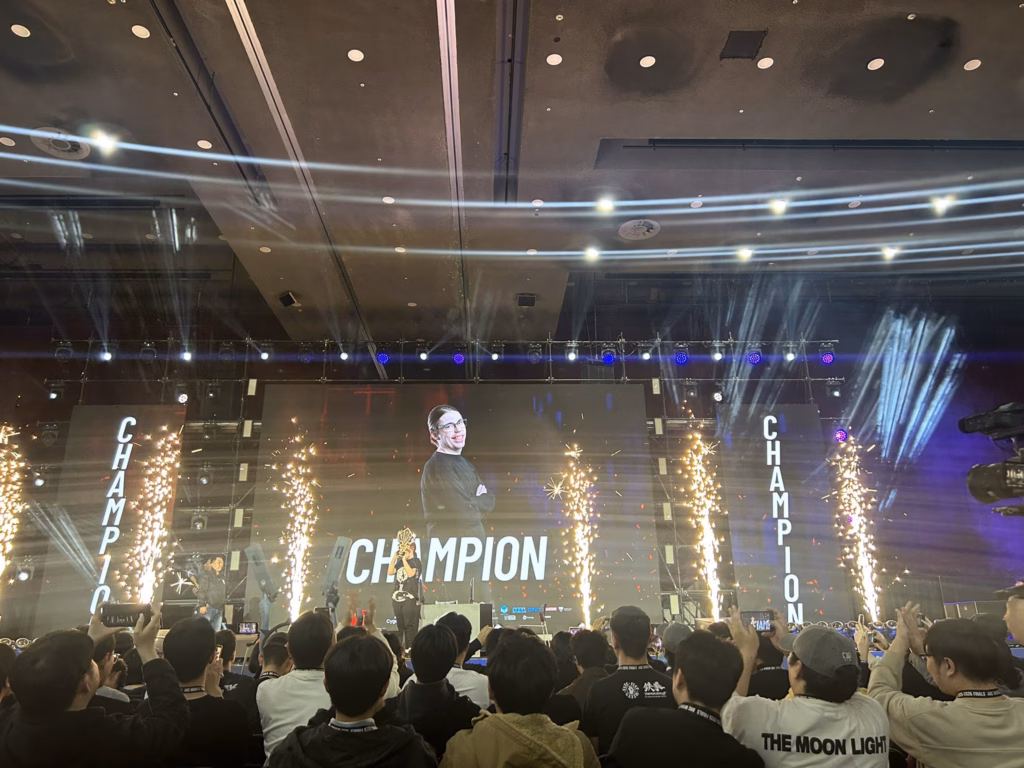 It’s a photo of the fighting games player Jack who won the Guilty Gear Strive tournament at the AWT 2025-2026 Finals - standing on the stage with the trophy, view from the audience.